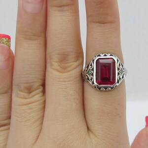 May include: A silver ring with a large, rectangular, red gemstone surrounded by a filigree design. Two smaller green gemstones are set on either side of the main stone.