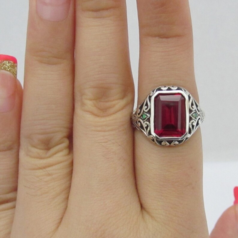 May include: A silver ring with a large, rectangular, red gemstone surrounded by a filigree design. Two smaller green gemstones are set on either side of the main stone.