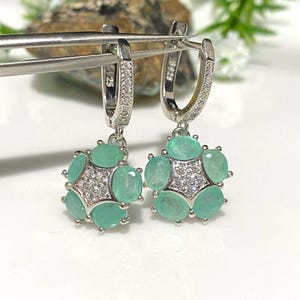 May include: A pair of silver-toned hoop earrings with a floral design. Each earring features a cluster of oval-cut, light green gemstones surrounding a central starburst of small, clear stones. The earrings are held by a silver tool.