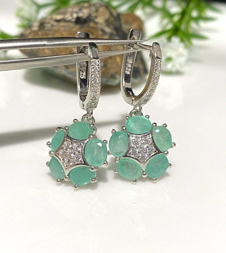 May include: A pair of silver-toned hoop earrings with a floral design. Each earring features a cluster of oval-cut, light green gemstones surrounding a central starburst of small, clear stones. The earrings are held by a silver tool.