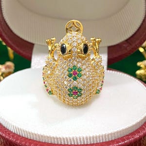 Op de afbeelding: Een gouden kikker hanger met een zwart emaille lichaam en groene en rode edelsteen accenten. De hanger is bedekt met heldere zirkonia stenen.