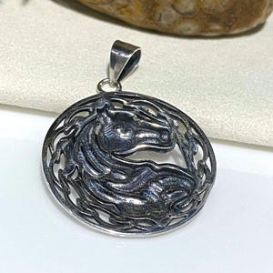 May include: A silver pendant featuring a detailed, stylized horse head design within a circular frame. The horse's head is facing right and the frame is made of intricate, interwoven lines.