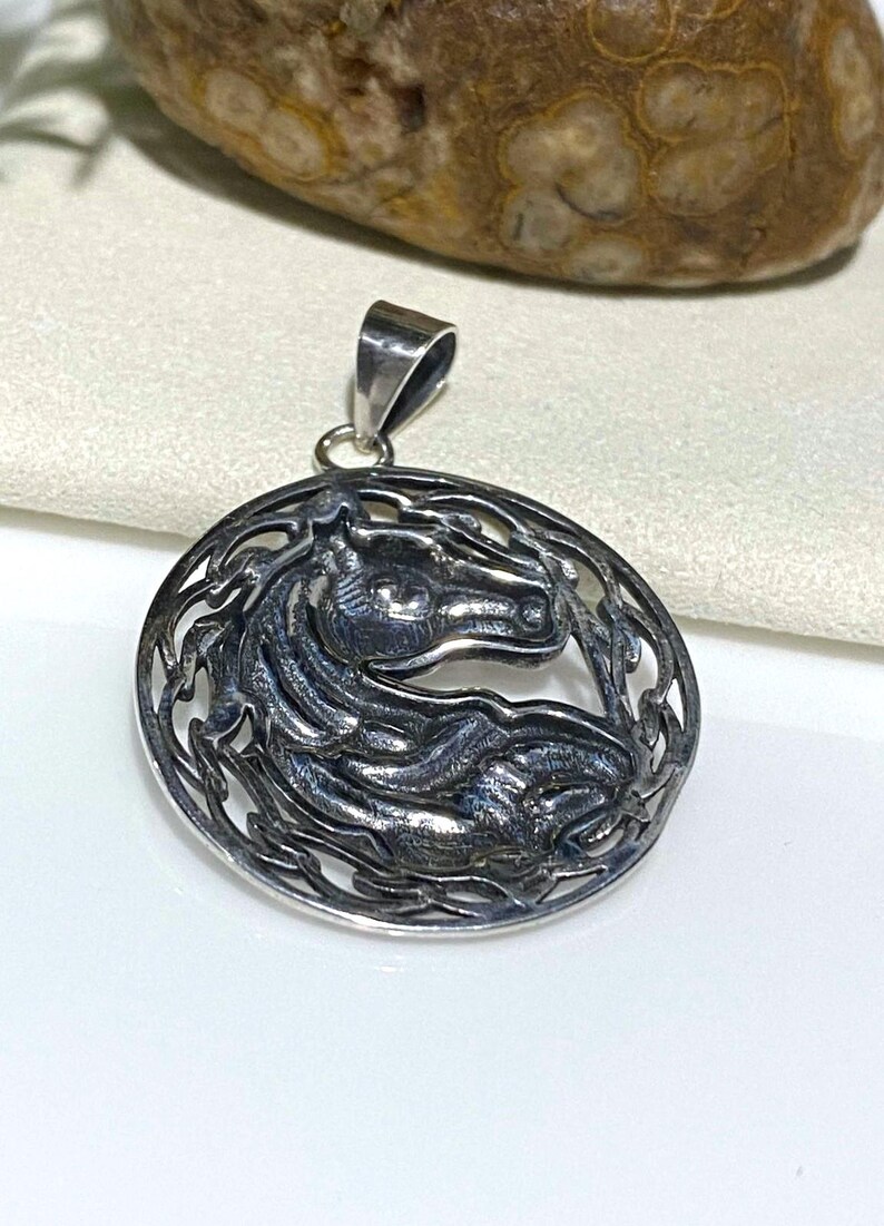 May include: A silver pendant featuring a detailed, stylized horse head design within a circular frame. The horse's head is facing right and the frame is made of intricate, interwoven lines.