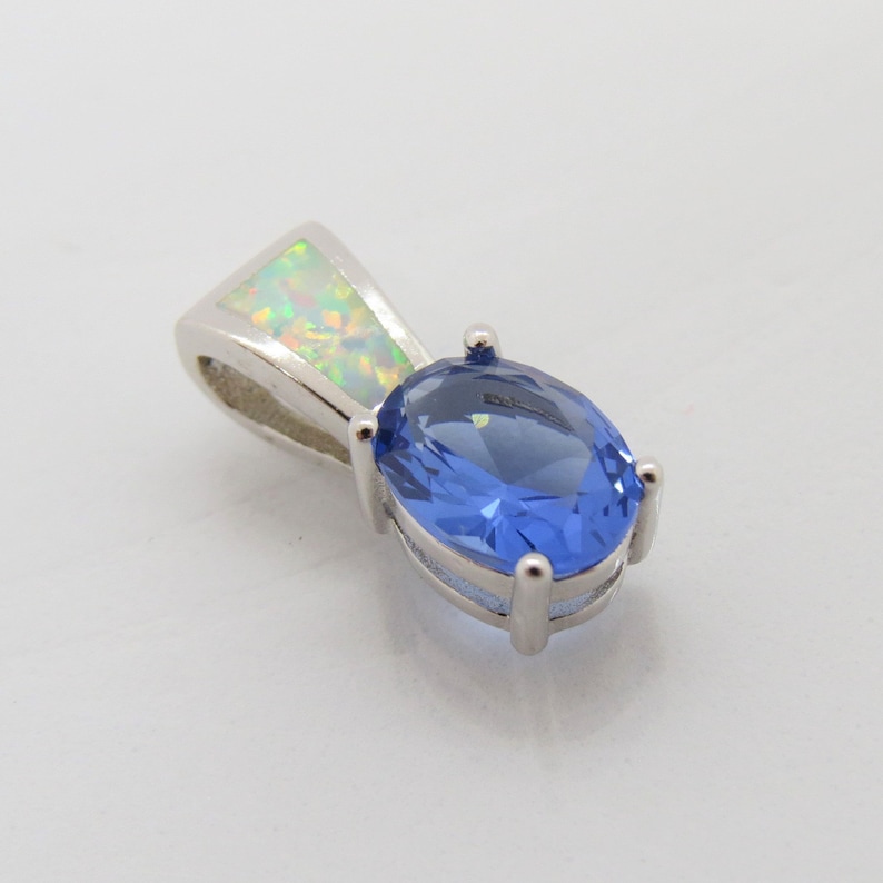 Op de afbeelding: Een zilveren hanger met een ovale, blauwe edelsteen en een driehoekige opaal. De hanger heeft een gepolijste afwerking en een klassiek ontwerp. De opaal toont een kleurenspel. Sieraad.