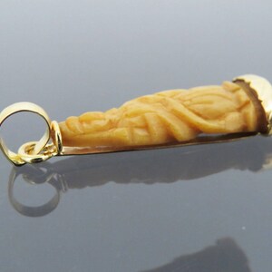 May include: A gold pendant featuring a carved figure with a detailed design. The pendant has a warm, light brown color with gold accents. The pendant is designed with a loop for a chain.