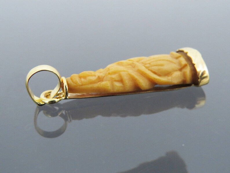 May include: A gold pendant featuring a carved figure with a detailed design. The pendant has a warm, light brown color with gold accents. The pendant is designed with a loop for a chain.