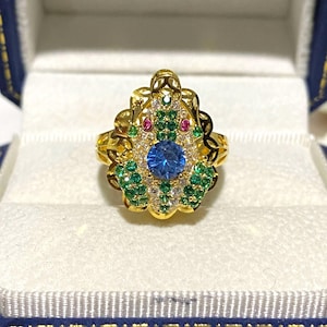 Op de afbeelding: Een gouden ring met een blauwe edelsteen omringd door kleinere groene en witte edelstenen. De ring heeft een delicaat, ingewikkeld ontwerp.