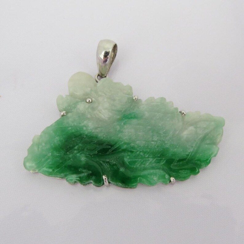 Pode incluir: Um pingente de jade verde e branco esculpido em forma de folha, engastado em prata com uma argola de prata.
