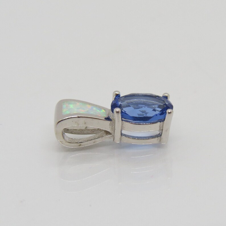 Op de afbeelding: Een zilveren hanger met een ovale, blauwe edelsteen en een kleinere, witte opaalsteen. De blauwe steen is gezet in een vier-klauwzetting. De hanger heeft een lus voor een ketting.