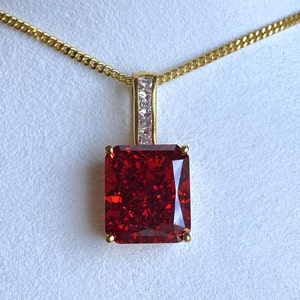 May include: A gold chain necklace with a red gemstone pendant. The pendant is a square cut with a clear stone border.