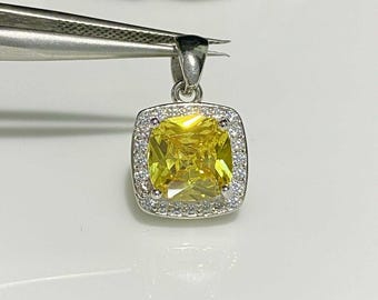 May include: A square, yellow gemstone pendant with a halo of small, clear stones set in a silver-colored metal. The pendant is held by tweezers against a white background. The gemstone is a vibrant, light yellow color.