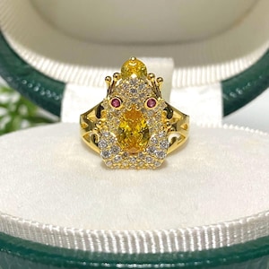 以下が含まれることがあります： 金色のカエルデザインのリング。カエルの体は大きな楕円形の黄色の宝石で、小さな赤と白の宝石が目を形成し、体を囲んでいます。リングには複雑なディテールが施されています。