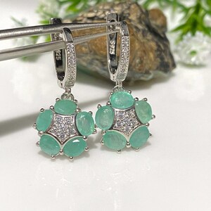 May include: A pair of silver earrings with a floral design. Each earring features a cluster of oval-cut, light green gemstones surrounding a central cluster of small, clear stones. The earrings are held by a silver clasp with small, clear stones.