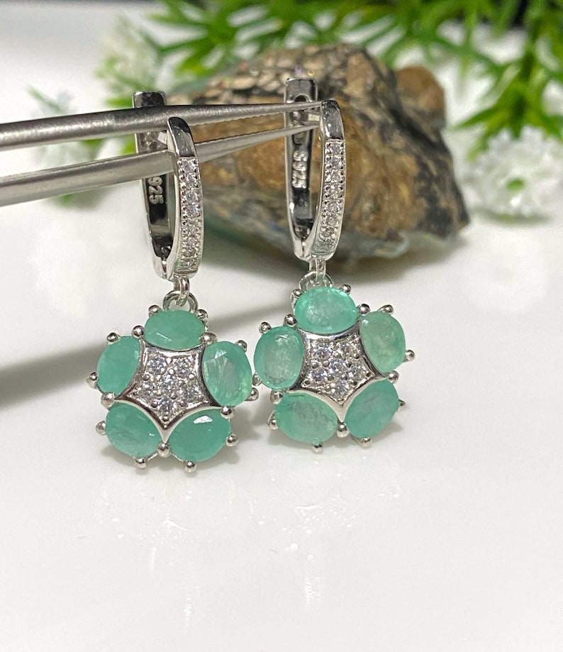May include: A pair of silver earrings with a floral design. Each earring features a cluster of oval-cut, light green gemstones surrounding a central cluster of small, clear stones. The earrings are held by a silver clasp with small, clear stones.