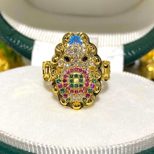 May include: A gold-colored ring with an ornate design featuring a central pattern of colorful gemstones. The ring has a large, detailed face with a blue stone at the top and a variety of other colored stones.