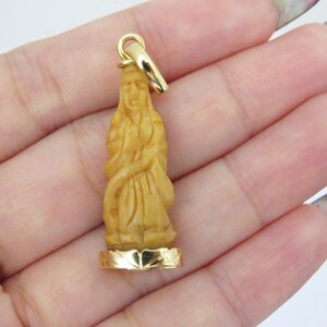 May include: A carved wooden pendant in the shape of a figure, with a gold-colored bail and base. The figure is light brown and detailed, with a flowing garment. The gold base has a decorative edge.