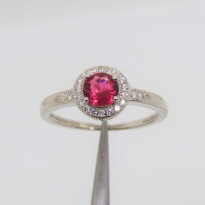 May include: A silver ring with a red gemstone surrounded by a halo of smaller white gemstones.