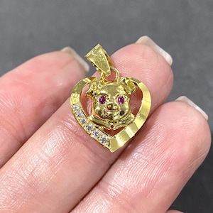 Puede incluir: Colgante dorado con la cabeza de un cerdo con ojos rojos, enmarcada en un corazón. El corazón está adornado con pequeñas gemas transparentes. El colgante tiene un lazo para una cadena.