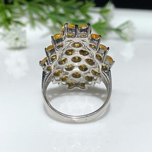 May include: A silver ring with a cluster of yellow gemstones set in a flower-like design. The ring is shown from the back, revealing the band and the setting.