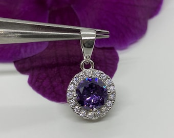 May include: A silver pendant with a round, purple gemstone surrounded by a halo of smaller, clear gemstones.