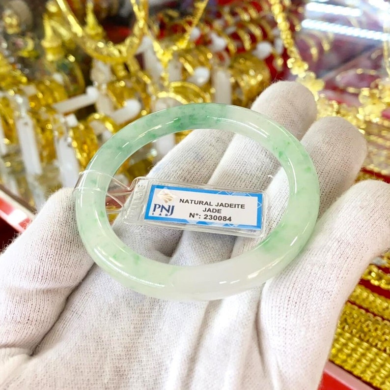 May include: A green jadeite jade bangle bracelet with a white background. The bracelet has a tag attached that reads "NATURAL JADEITE JADE N.: 230084".
