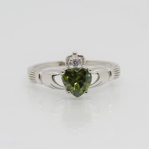 May include: A silver Celtic Claddagh ring featuring a green heart-shaped gemstone and a small clear stone on the crown.