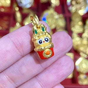 May include: A gold-colored pendant in the shape of a rabbit in a red cup. The rabbit has green accents on its ears and a cute face. The cup has an orange emblem. The pendant is held in a person's hand.