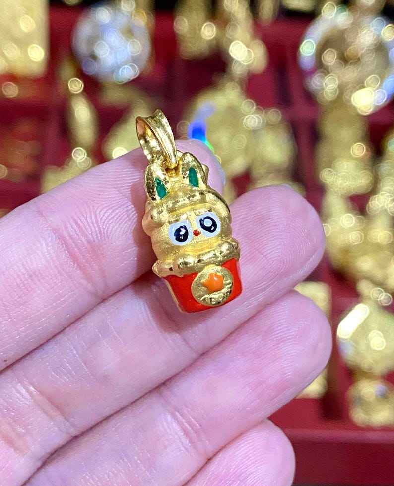 May include: A gold-colored pendant in the shape of a rabbit in a red cup. The rabbit has green accents on its ears and a cute face. The cup has an orange emblem. The pendant is held in a person's hand.