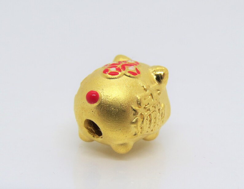 May include: A gold-colored ceramic pig figurine with red accents. The pig has a hole in the back, suggesting it may be a piggy bank or a charm.
