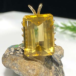 May include: A gold pendant featuring a large, faceted, yellow gemstone with a bezel setting. The gemstone is rectangular in shape and has a smooth, polished finish.