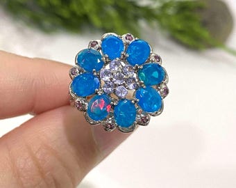 May include: A silver ring featuring a floral design with blue oval-cut gemstones. The ring's center has smaller purple gemstones, and the outer edge is accented with small pink gemstones. The ring is held by a hand.