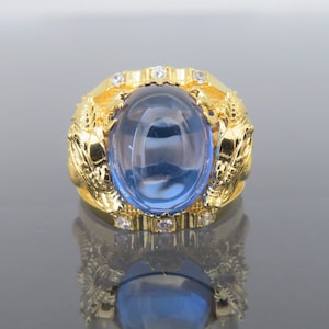 May include: A gold ring featuring a large blue oval gemstone surrounded by smaller clear gemstones. The ring is adorned with a detailed design of two dragons on either side of the gemstone.