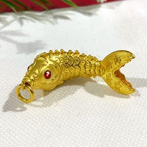 Puede incluir: Un amuleto de pez de color dorado con ojos de piedra roja. El amuleto est&aacute; detallado con escamas y tiene un bucle para sujetarlo a un collar o pulsera.