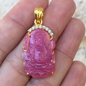 May include: Gold pendant featuring a pink carved gemstone with a seated Buddha figure. The pendant is accented with a halo of clear cubic zirconia stones.