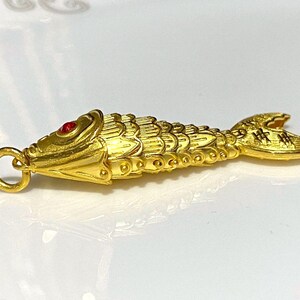 Puede incluir: Un amuleto de pez de color dorado con un ojo de piedra roja. El pez est&aacute; detallado con escamas y tiene un bucle para sujetarlo a una cadena o pulsera.