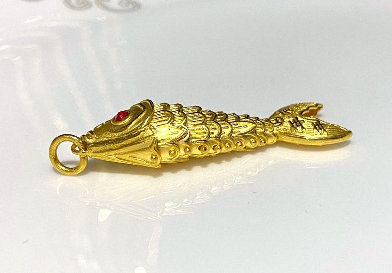 Puede incluir: Un amuleto de pez de color dorado con un ojo de piedra roja. El pez est&aacute; detallado con escamas y tiene un bucle para sujetarlo a una cadena o pulsera.