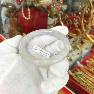 Pode incluir: Um bracelete de jadeíta branco com um tom verde claro. O bracelete é apresentado em um pano branco e tem uma etiqueta que diz "PNJ NATURAL JADEITE No: 131756".