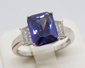 Op de afbeelding: Een zilveren ring met een grote, rechthoekige, donkerblauwe edelsteen omringd door kleinere, heldere edelstenen.