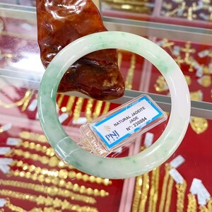 May include: A white jadeite bangle bracelet with green flecks. The bracelet is on display with a tag that reads "NATURAL JADEITE JADE N&deg;. 230084" and a red background.