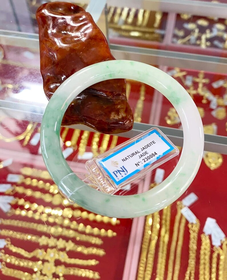 May include: A white jadeite bangle bracelet with green flecks. The bracelet is on display with a tag that reads "NATURAL JADEITE JADE N&deg;. 230084" and a red background.