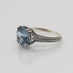 May include: A silver ring featuring a light blue gemstone set in a filigree design. The band is decorated with a repeating pattern of small leaves.