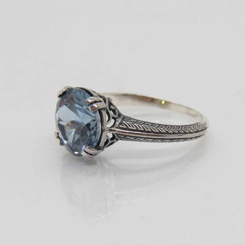 May include: A silver ring featuring a light blue gemstone set in a filigree design. The band is decorated with a repeating pattern of small leaves.