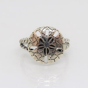 May include: Silver ring with a detailed sand dollar design. The band is twisted and has a textured finish.