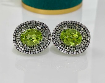 May include: A pair of oval-shaped peridot gemstone stud earrings. Each earring features a vibrant green peridot center stone surrounded by a halo of smaller, sparkling gemstones. The earrings are set in a silver-toned metal.