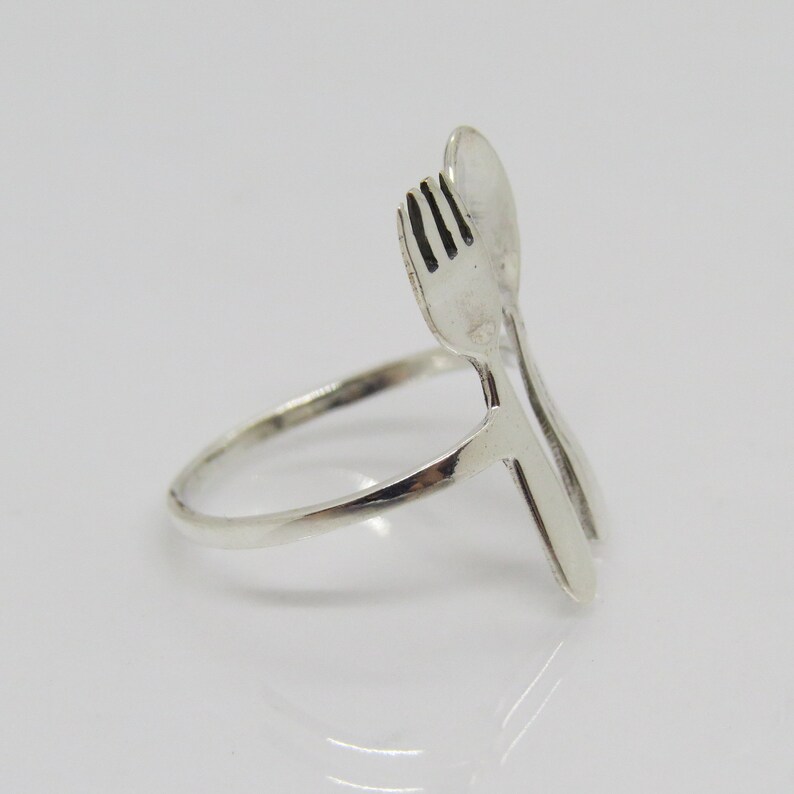 Puede incluir: Un anillo de plata con un dise&ntilde;o de tenedor y cuchara. El tenedor y la cuchara est&aacute;n entrelazados y forman la banda del anillo.
