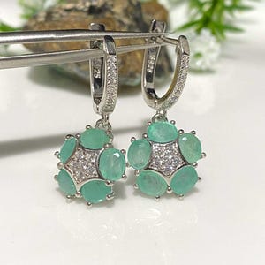 May include: A pair of silver-toned hoop earrings. Each earring features a cluster of oval-cut, light green gemstones surrounding a central starburst of smaller, clear stones. The earrings are displayed on a white surface.