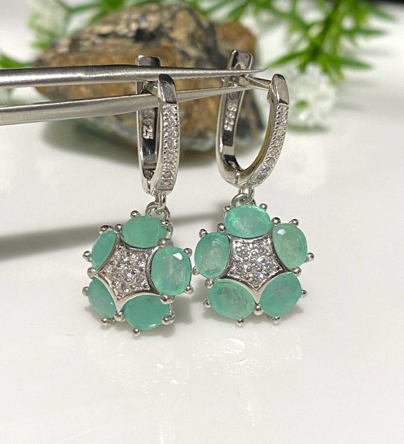 May include: A pair of silver-toned hoop earrings. Each earring features a cluster of oval-cut, light green gemstones surrounding a central starburst of smaller, clear stones. The earrings are displayed on a white surface.