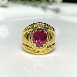 May include: A gold ring with a large, oval-shaped, dark red gemstone. The ring has a band with an inscription around the top.