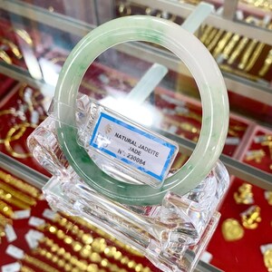 May include: A green jadeite jade bangle bracelet on a clear display stand. The tag reads "NATURAL JADEITE JADE N&deg; 230084".