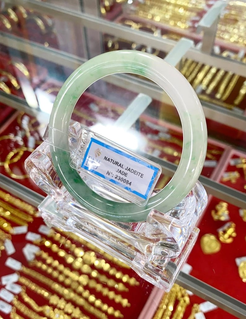 May include: A green jadeite jade bangle bracelet on a clear display stand. The tag reads "NATURAL JADEITE JADE N&deg; 230084".